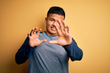 Sarı arka planda dikilen sıradan tişörtlü genç, yakışıklı Latin adam korku ifadesinden korkuyor ve dehşete düşüyor. El hareketlerini durdur, şok içinde bağır. Panik konsepti.