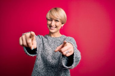 Modern kısa saçlı, pembe arka planda rahat süveter giyen genç sarışın kadın seni ve kamerayı parmaklarıyla gösteriyor, gülümsüyor ve neşeli.