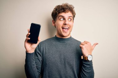 Sarışın, kıvırcık saçlı, akıllı telefon gösteren genç bir adam izole edilmiş beyaz arka plan üzerinde duruyor ve gülümseyen bir gülümsemeyle baş parmağıyla onu işaret ediyor.
