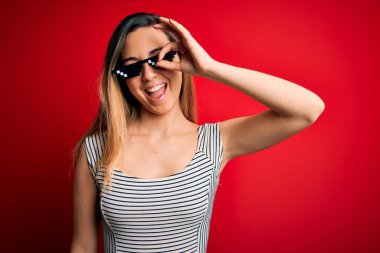 Genç, güzel esmer, komik, cani hayat gözlüğü takan bir kadın. Kırmızı arka planda güneş gözlüğü takıyor. El hareketleriyle gülümsüyor..