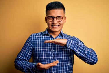 Sarı arka plan üzerine gündelik tişört ve gözlük takan genç, yakışıklı Latin bir adam. Elleri büyük ve büyük boy işareti, ölçü sembolü gösteriyor. Kameraya bakıp gülümsüyor. Ölçüm konsepti.