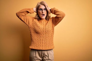 Orta yaşlı güzel sarışın kadın sarı arka planda gözlük ve süveter takıyor. Çılgına dönmüş, elleri kafasında korkmuş ve ağzı açık şok geçirmekten korkmuş.