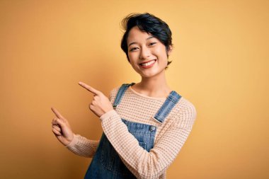 Genç, güzel Asyalı kız izole edilmiş sarı arka planda günlük kot pantolon giyiyor gülümsüyor ve kameraya bakıyor iki eliyle ve parmaklarıyla yan tarafı işaret ediyor..