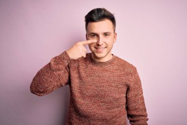 Pembe izole edilmiş arka plan üzerine sıradan bir kazak giyen genç, yakışıklı, beyaz bir adam. El ve burunla işaret ediyor, neşeli gülümsüyor. Güzellik konsepti