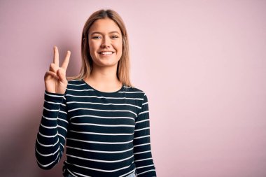 Pembe izole edilmiş arka plan üzerine sıradan çizgili süveter giyen genç ve güzel sarışın kadın kameraya gülümseyip zafer işareti yapıyor. İki numara..