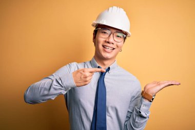 Güvenlik kaskı takan ve sarı arka planda kravat takan genç yakışıklı mimar kameraya hayret ediyor ve gülümsüyor..