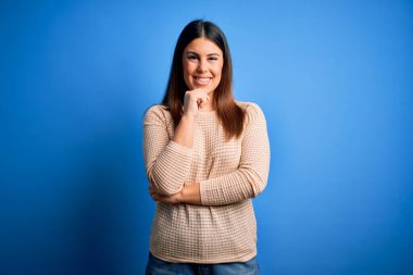 Mavi arka plan üzerine sıradan süveter giyen genç ve güzel bir kadın kameraya kollarını kavuşturup gülümseyerek ve çenesini kaldırarak bakıyor. Olumlu düşünüyorum..