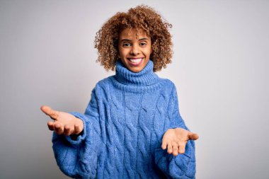 Güzel Afro-Amerikan kadın balıkçı yaka kazak giyip beyaz arka planda gülümsüyor. Kollarını açıp gülümsüyor. Dostça hoş geldin, olumlu ve kendinden emin selamlar.