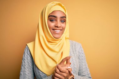 Genç ve güzel Afro-Amerikalı kız izole edilmiş sarı arka planda Müslüman başörtüsü takıyor ellerini birleştirip gülümseyerek gevşemiş ve neşeli gülümsüyor. Başarı ve iyimser
