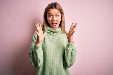 Young beautiful blonde woman wearing winter wool sweater over pink isolated background celebrating crazy and amazed for success with arms raised and open eyes screaming excited. Winner concept