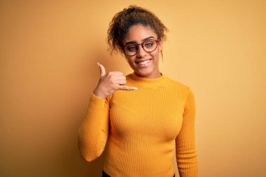 Sarı arka planda süveter ve gözlüklerle gülümseyen, el ve parmaklarıyla telefonla konuşan güzel Afro-Amerikan kızı. İletişim kavramları.