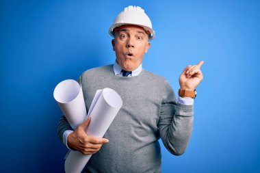 Güvenlik kaskı takan, orta yaşlı, yakışıklı, gri saçlı bir mimar elinde planları olan, parmaklarını yana doğru gösteren, ağzı açık, şaşırmış bir mimar..