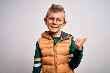 Mavi gözlü küçük beyaz çocuk kışlık palto giyiyor ve güzel gözlükler takıyor. Gülümsüyor, gülümsüyor ve başparmağıyla yana bakıyor..