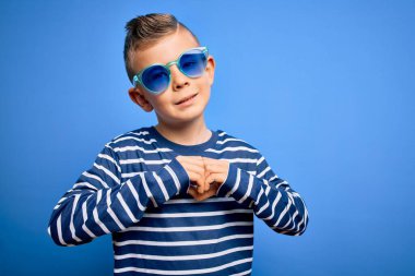 Mavi gözlü küçük beyaz çocuk mavi arka planda güneş gözlüğü takıyor. Aşkla gülümsüyor. Elleriyle kalp sembolü ve şekli gösteriyor. Romantik konsept.