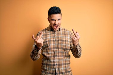 Young handsome man wearing casual shirt standing over isolated yellow background Showing middle finger doing fuck you bad expression, provocation and rude attitude. Screaming excited
