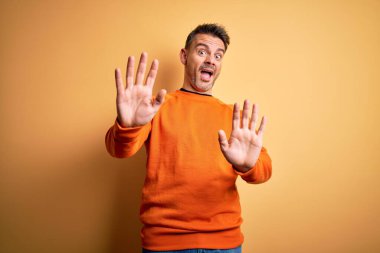 Sarı arka plan üzerinde duran turuncu renkli süveter giyen genç ve yakışıklı adam korku ifadesinden korkmuş ve dehşete düşmüş. El hareketlerini durdur, şok içinde bağır. Panik konsepti.