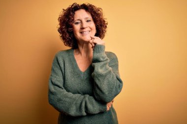 Orta yaşlı, kıvırcık saçlı, rahat sarı arka planda süveter giyen güzel bir kadın kameraya kollarını kavuşturup gülümseyen ve çenesini kaldıran bir şekilde bakıyor. Olumlu düşünüyorum..