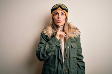 Genç esmer kayakçı kadın kar kıyafetleri ve kayak gözlüğü takmış beyaz arka planda şüpheye odaklanmış, çenesinde parmakları olan ve kafasını kaldırıp,