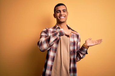 Sarı arka planda dikilen ve kameraya gülümseyen gündelik gömlekli Afro-Amerikan genç adam elini uzatıp parmağıyla işaret ediyor..