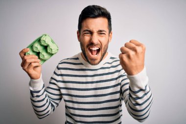 Sakallı, sakallı, beyaz arka planda taze yumurta kutusuyla gezen yakışıklı genç bir adam sinirlenmiş ve sinirlenmiş bir şekilde bağırıyor.