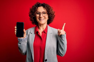 Orta yaşlı kıvırcık kadın elinde akıllı telefon ile izole edilmiş kırmızı arka plan üzerinde bir fikir ya da soru işareti ile şaşırmış bir numara, mutlu yüzlü, bir numara