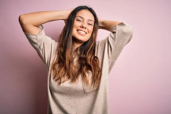 Young beautiful brunette woman wearing casual sweater standing over pink background relaxing and stretching, arms and hands behind head and neck smiling happy