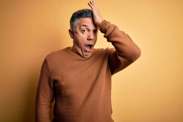Middle age handsome grey-haired man wearing casual sweater over yellow background surprised with hand on head for mistake, remember error. Forgot, bad memory concept.