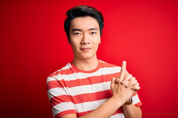 Young handsome chinese man wearing casual striped t-shirt standing over red background Holding symbolic gun with hand gesture, playing killing shooting weapons, angry face