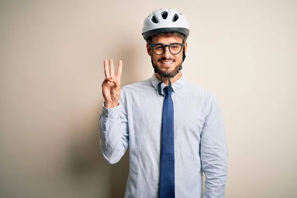 Young businessman wearing glasses and bike helmet standing over isolated white bakground showing and pointing up with fingers number three while smiling confident and happy.