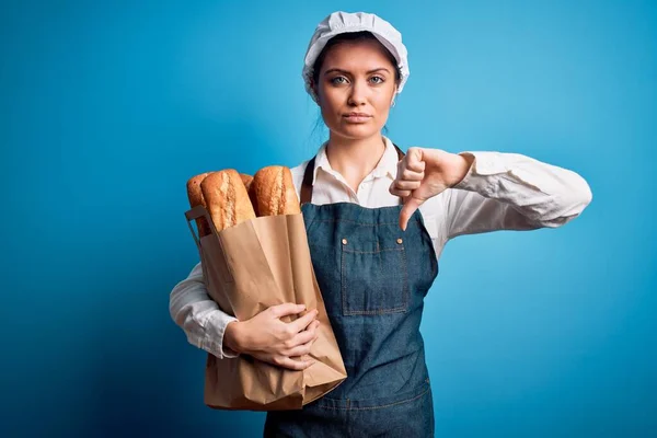 年轻美丽的面包师女人 蓝眼睛 手里拿着装有面包的纸袋 满脸怒容 消极的标志 对拇指朝下表示不喜欢 拒绝的观念 — 图库照片