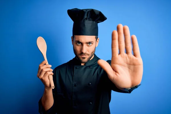 年轻的炊事员 留着胡子 身穿制服 蓝色背景上拿着木勺 张开手 做停止手势 表情严肃自信 防守姿势 — 图库照片