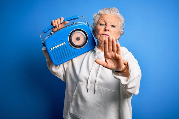 年长而美丽的女人手持老式收音机 站在孤立的蓝色背景上 张开双手 做着手势 严肃而自信地表达着防守的姿态 — 图库照片