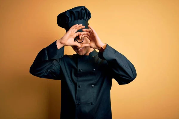 Young brazilian chef man wearing cooker uniform and hat over isolated yellow background Doing heart shape with hand and fingers smiling looking through sign