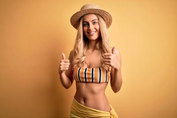 Young beautiful blonde woman on vacation wearing bikini and hat over yellow background success sign doing positive gesture with hand, thumbs up smiling and happy. Cheerful expression and winner gesture.