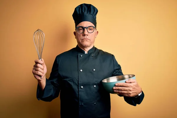 Middle age handsome grey-haired chef man wearing cooker uniform and hat using whisk with a confident expression on smart face thinking serious