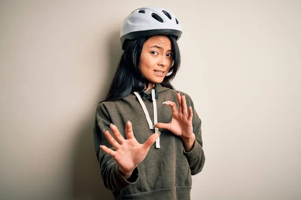 Young beautiful chinese woman wearing bike helmet over isolated white background disgusted expression, displeased and fearful doing disgust face because aversion reaction. With hands raised