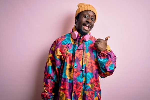 Young handsome african american man wearing colorful coat and cap over pink background smiling with happy face looking and pointing to the side with thumb up.