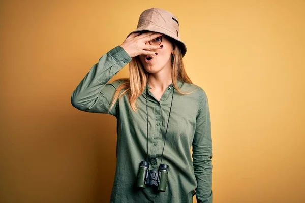 Beautiful blonde explorer woman with blue eyes wearing hat and glasses using binoculars peeking in shock covering face and eyes with hand, looking through fingers with embarrassed expression.