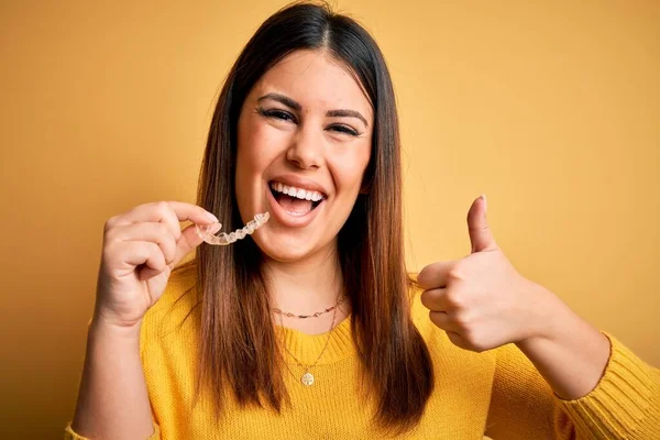 年轻美丽的女人带着矫正牙齿的矫形器在黄色的背景上快乐的笑着做着好的手势 用手指竖起大拇指 很好的标志 — 图库照片