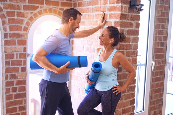 Middle age beautiful sporty couple smiling happy. Standing with smile on face holding mat and speaking at gym