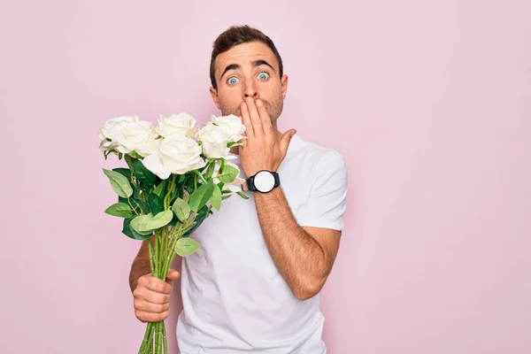Young handsome man with blue eyes holding bouquet of flowers over pink background cover mouth with hand shocked with shame for mistake, expression of fear, scared in silence, secret concept