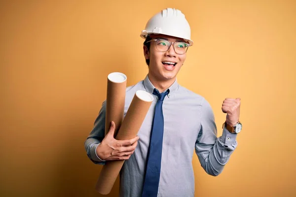 Young handsome chinese architect man wearing glasses and safety helmet holding blueprints pointing and showing with thumb up to the side with happy face smiling
