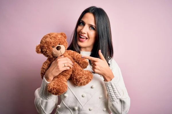 Young brunette woman with blue eyes hugging teddy bear stuffed animal over pink background very happy pointing with hand and finger