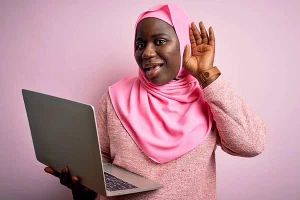 African american plus size woman wearing muslim hijab using laptop over pink background smiling with hand over ear listening an hearing to rumor or gossip. Deafness concept.