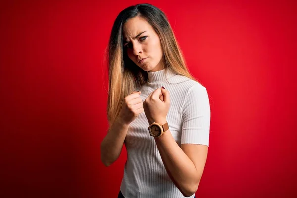 Beautiful blonde woman with blue eyes wearing casual white t-shirt over red background Ready to fight with fist defense gesture, angry and upset face, afraid of problem