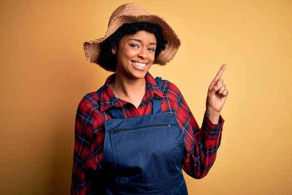 Young African American afro farmer woman with curly hair wearing apron and hat with a big smile on face, pointing with hand and finger to the side looking at the camera.