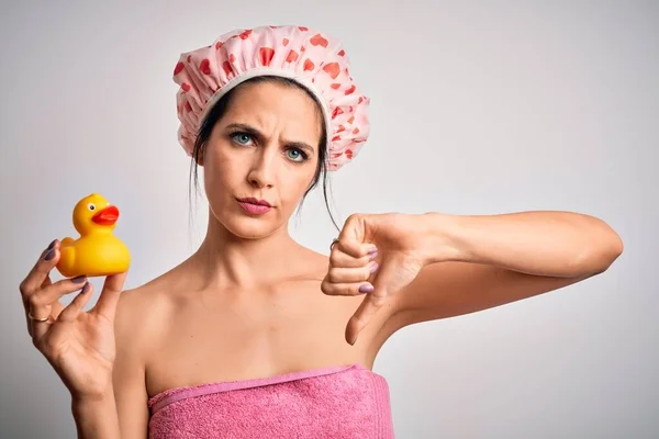 Young brunette woman with blue eyes wearing bath towel and shower cap holding duck toy with angry face, negative sign showing dislike with thumbs down, rejection concept