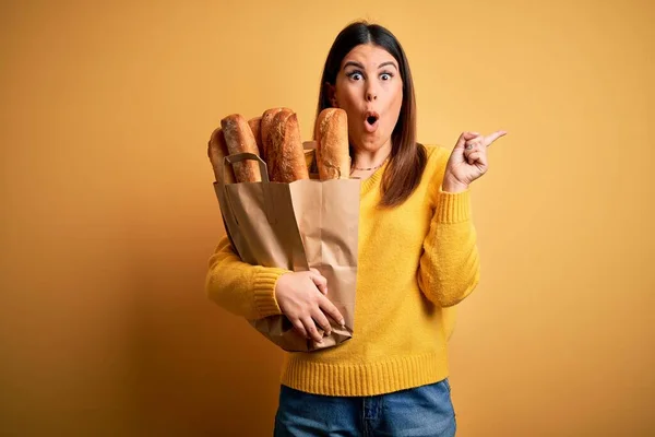 年轻美丽的女人 提着一袋黄色背景的新鲜健康的面包 用手指指向侧面 张开嘴 惊讶的表情 — 图库照片
