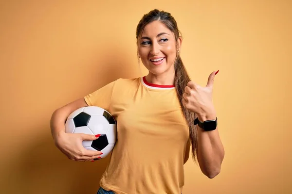 美しいブルネットのプレーヤーの女性プレイサッカーボールを使用して黄色の背景ポインティングと表示上の親指で側まで幸せな顔笑顔 — ストック写真