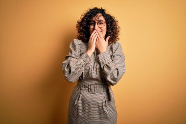 Güzel bir Arap iş kadını elbise ve gözlük takıyor sarı arka planda gülüyor ve utanıyor kıkırdıyor ağzı ellerle, dedikoduyla ve skandalla örtüyor.